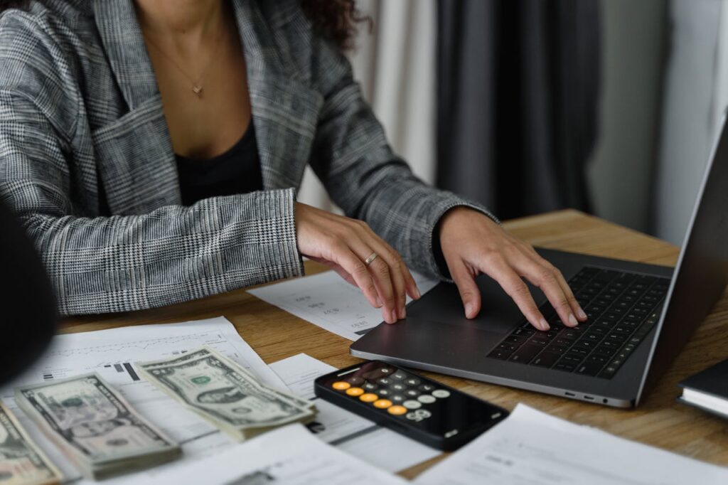 A woman is at her desk with her laptop, US dollar bills, and calculator consolidating her debt.