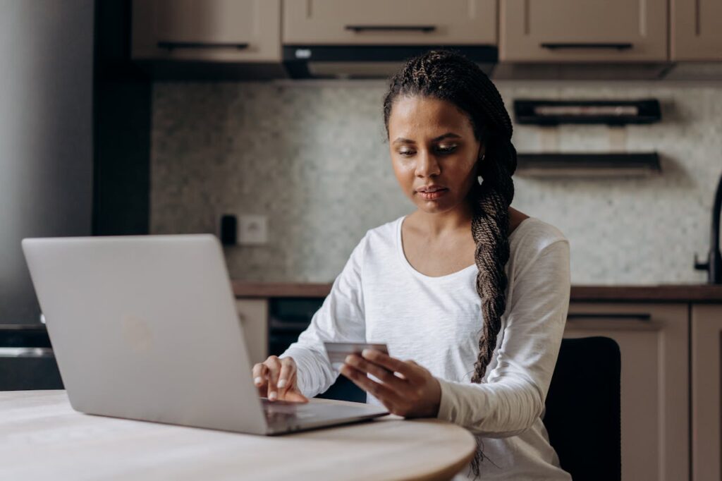 A woman is sitting at her kitchen table with her laptop and credit card, thinking of whether to use it for debt consolidation.