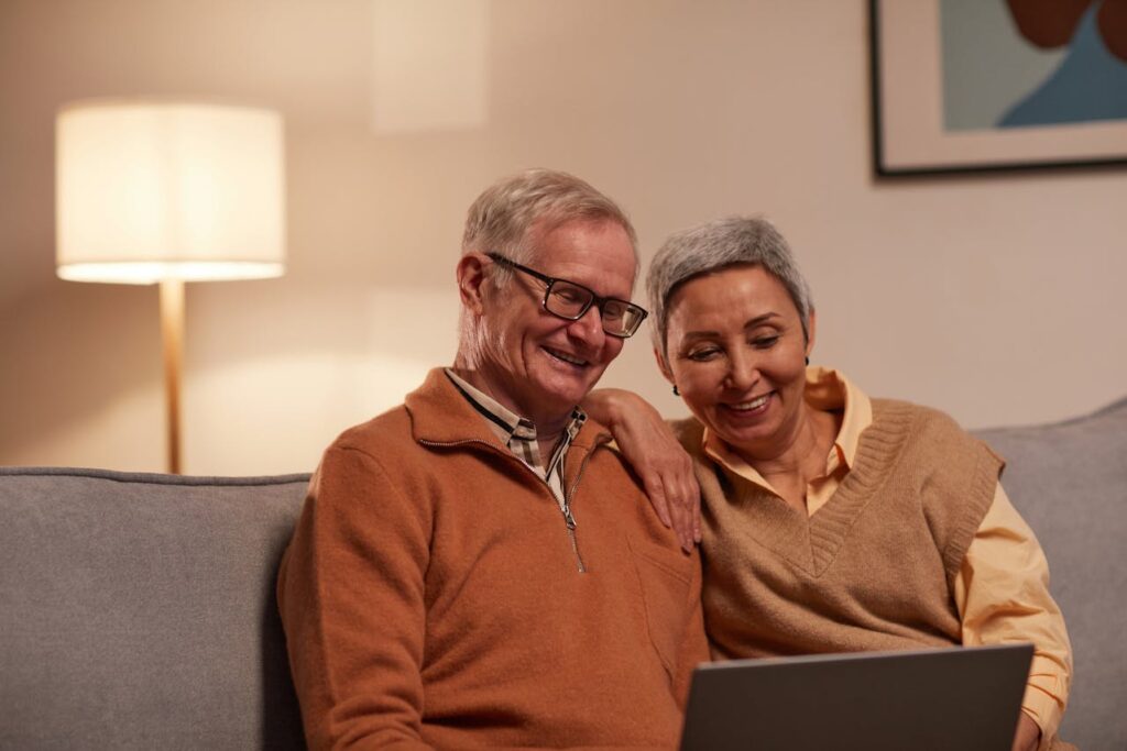 An elderly couple is sitting on a couch and looking at a laptop screen to review their options for mortgages for seniors.