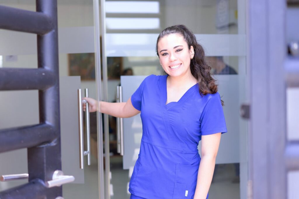 A nurse in blue scrubs is smiling while holding a door open. 