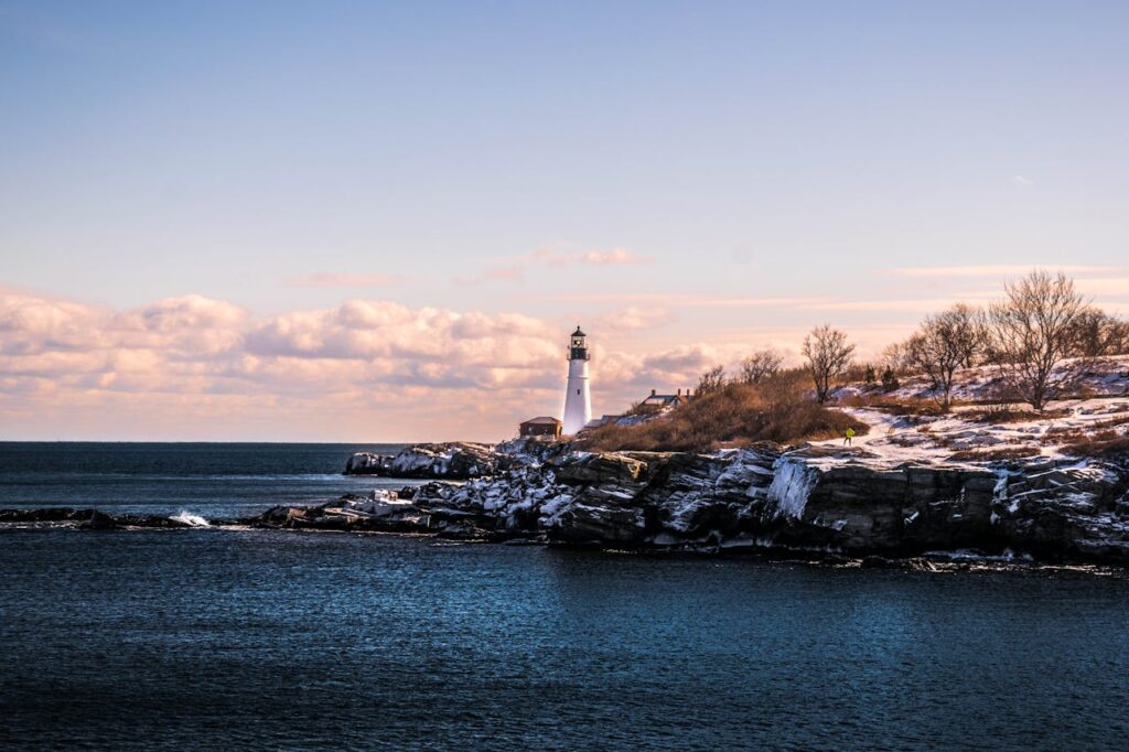 A lighthouse on the coast in Maine with snow covering the hills beside it. 