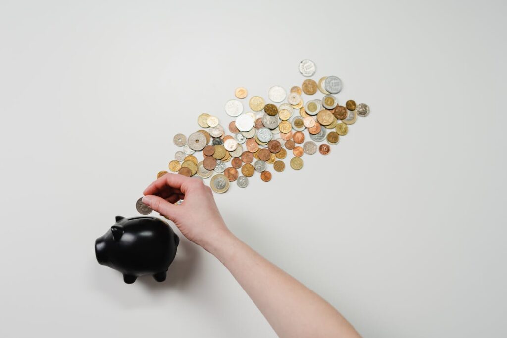 A piggy bank with various coins coming out of it while a hand is inserting another coin. 