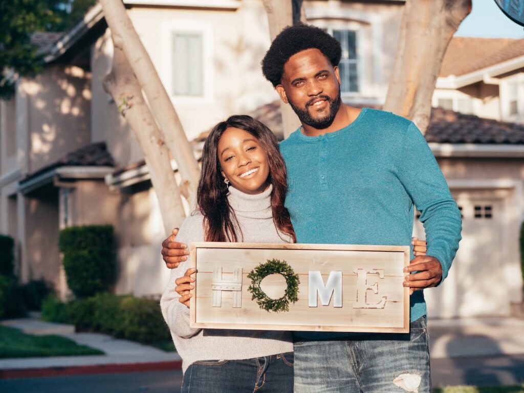 A young couple is holding a sign in front of their house that says "home." 